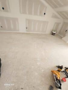 A room under construction with unfinished drywall, exposed outlets, and a bare floor. Construction tools and supplies are scattered in the lower right corner.
